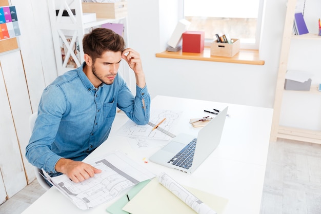 Konzentrierter junger Geschäftsmann, der mit Dokumenten arbeitet, während er mit Laptop im Büro sitzt