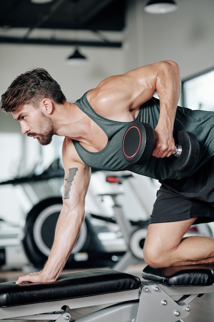 Konzentrierter junger hübscher erwachsener Mann, der einarmige Hantelreihen auf Bank im modernen Fitnessstudio tut