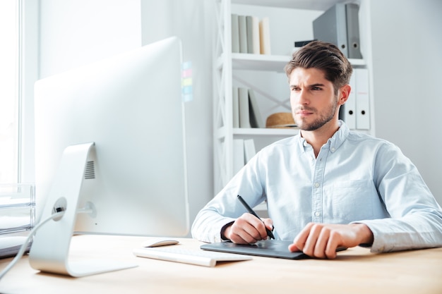 Konzentrierter junger Mann Designer, der am Arbeitsplatz mit Computer und Grafiktablett arbeitet tablet