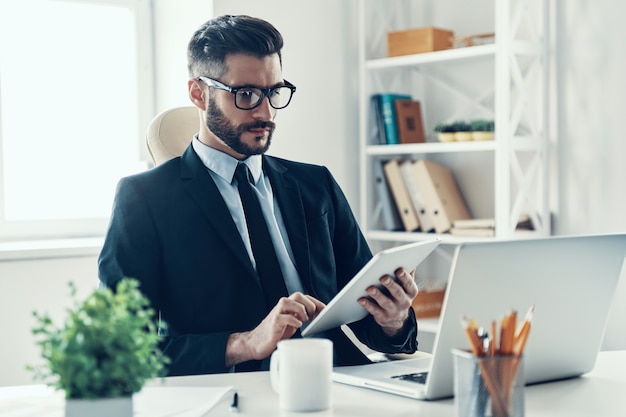 Konzentrierter junger Mann im eleganten Business-Anzug, der mit digitalem Tablet arbeitet, während er im Büro sitzt