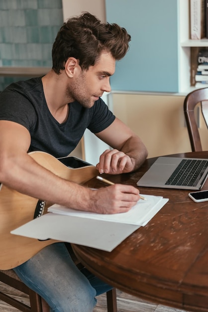 Konzentrierter junger Mann mit Gitarre, der Notizen schreibt, während er auf den Laptop schaut