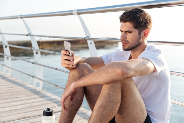 Konzentrierter junger Sportler, der auf Pier sitzt und Smartphone benutzt