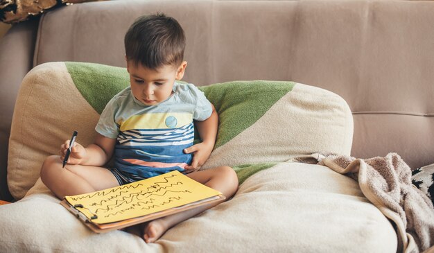 Konzentrierter kleiner Junge, der auf einem gelben Papier unter Verwendung eines Markers zeichnet, posiert auf Bett