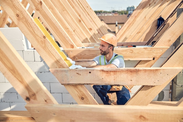 Konzentrierter Mann mit Schutzhelm, der eine Wasserwaage an eine diagonale Holzschiene hält