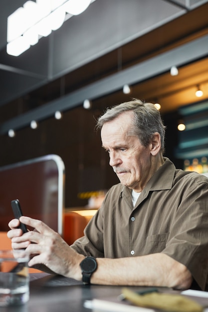 Konzentrierter reifer Mann mit Schnurrbart, der am Tisch sitzt und mobiles Internet auf dem Smartphone im Café mit kostenlosem WLAN nutzt