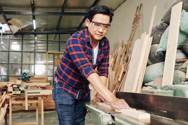 Konzentrierter reifer Zimmermann im karierten Hemd, der lange Holzbretter in zwei Teile sägt