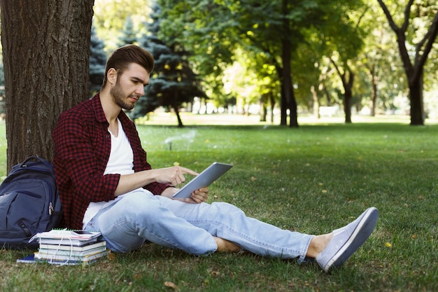 Konzentrierter Student, der sich im Park auf die Prüfung vorbereitet. Legerer Mann mit digitalem Tablet, sitzend auf Gras, Kopierraum. Technologie-, Kommunikations-, Bildungs- und Fernarbeitskonzept.