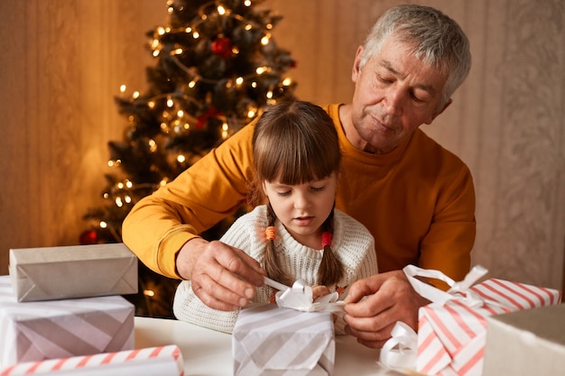 Konzentriertes kleines Mädchen mit Zöpfen, das einen weißen Pullover trägt, der mit ihrem Großvater am Tisch posiert und Geschenkkartons vorbereitet, um ihrer Familie zu Silvester zu gratulieren.