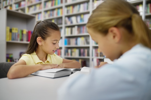 Konzentriertes schönes kaukasisches dunkelhaariges Mädchen und ihre blonde Klassenkameradin lesen Bücher in einer öffentlichen Bibliothek