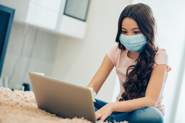 Konzentriertes Schulmädchen, das Hausaufgaben auf dem Laptop erledigt, während es mit einer medizinischen Maske im Gesicht auf dem Bett sitzt