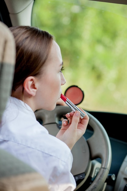 Konzept der Gefahr beim Fahren Junge Fahrerin, rothaarige Teenagerin, die ihre Lippen beim Auftragen von Make-up beim Autofahren malt