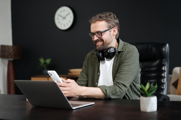 Foto konzept der kommunikation und technologie am arbeitsplatz europäischer mann, der sms auf dem mobiltelefon schreibt, während er im büro am laptop arbeitet multitasking-konzentration und nahtlose verbindung zwischen zwei geräten