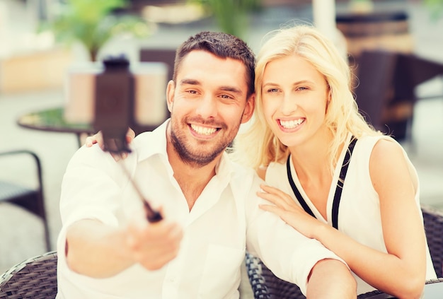 Konzept der Liebe, des Datums, der Technologie, der Menschen und der Beziehungen - glückliches glückliches Paar, das mit dem Smartphone auf dem Selfie-Stick im Straßencafé oder Restaurant der Stadt fotografiert