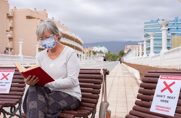 Konzept der sozialen Distanzierung. Ältere Frau, die allein auf Holzbank sitzt und ein Buch liest reading