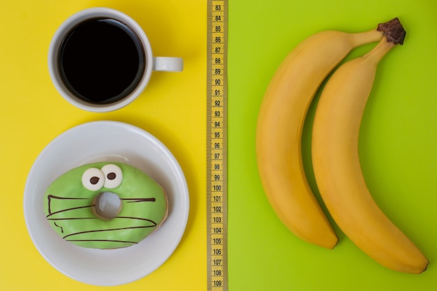 Konzept der ungesunden Ernährung und Snacks. Köstlicher Donut und Tasse starker Kaffee einzeln auf gelben und frischen leckeren Bananen einzeln auf grünem Hintergrund. Foto von oben