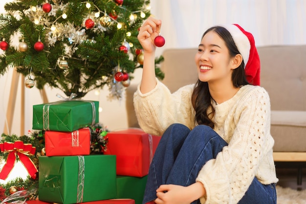 Konzept der Weihnachtsfeier Junge asiatische Frau mit rotem Ball zum Dekorieren im Weihnachtsbaum