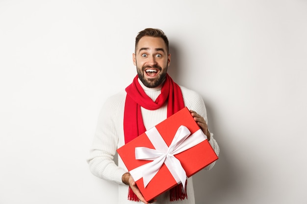 Konzept der Winterferien. Überraschter Mann, der ein Weihnachtsgeschenk erhält und erstaunt aussieht, eine rote verpackte Schachtel mit Geschenk hält und auf weißem Hintergrund steht