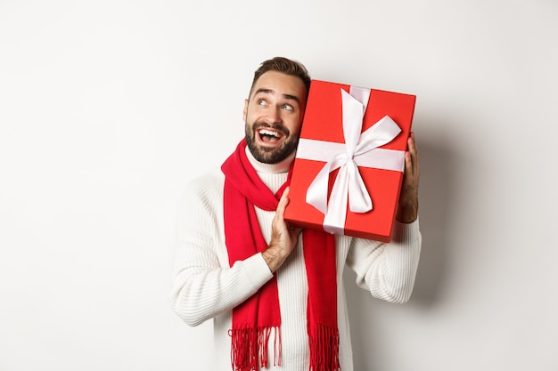 Konzept der Winterferien. Glücklicher Mann, der Geschenkbox schüttelt, um zu erraten, was drin ist, neugierig aussieht, Weihnachtsgeschenke erhält und auf weißem Hintergrund steht.