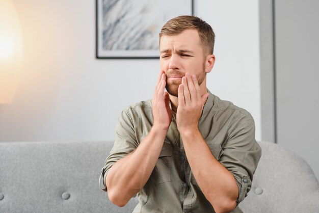 Konzept der Zahngesundheitsprobleme Mann, der unter starken Zahnschmerzen leidet Verärgerter männlicher Patient mit entzündetem Nervenzahnabszess oder Zahnempfindlichkeit, der auf dem Sofa sitzt und die Wange mit Schmerzensgrimasse berührt