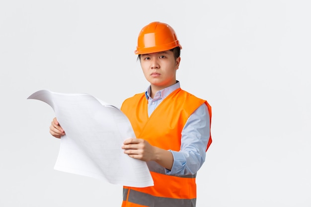 Konzept des Bausektors und der Industriearbeiter. Ernsthaft aussehender selbstbewusster asiatischer Bauleiter, Ingenieur mit Helm und reflektierender Jacke mit Blick auf die Blaupause, Überprüfung des Layouts, weißer Hintergrund.