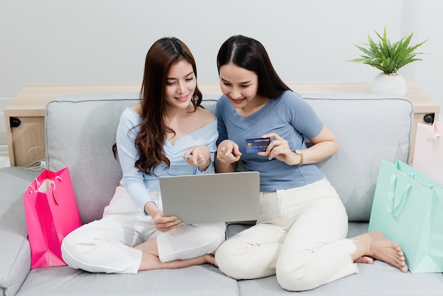 Foto konzept des online-shoppings von zu hause aus zwei schöne asiatische frauen verwenden eine kreditkarte, um ihren laptop-spaß auf dem sofa zu hause auszufüllen und papiertüten einzukaufen. neues normales online-geschäftsmodell