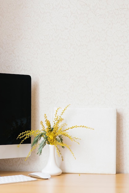 Konzept eines Business-Freelancer-Bloggers Der Laptop steht auf einem Holztisch an einer weißen Wand mit einer Mimose in einer Vase