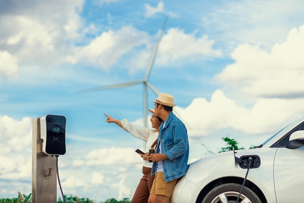 Konzept eines progressiven, glücklichen jungen Paares an einer Windkraftanlage mit Elektrofahrzeug