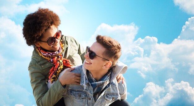 Konzept Freundschaft, Freizeit, Freiheit und Menschen - glückliches internationales Teenagerpaar in Schattierungen, das sich über blauem Himmel und Wolkenhintergrund amüsiert