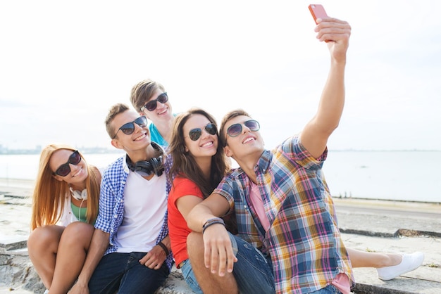 Konzept Freundschaft, Freizeit, Sommer, Technologie und Menschen - Gruppe glücklicher Freunde mit Smartphone, die sich im Freien selbst machen