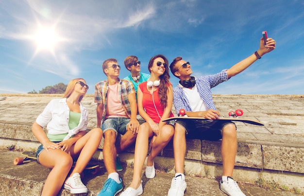 Konzept Freundschaft, Freizeit, Sommer, Technologie und Menschen - Gruppe lächelnder Freunde mit Skateboard und Smartphone, die sich im Freien selbst machen