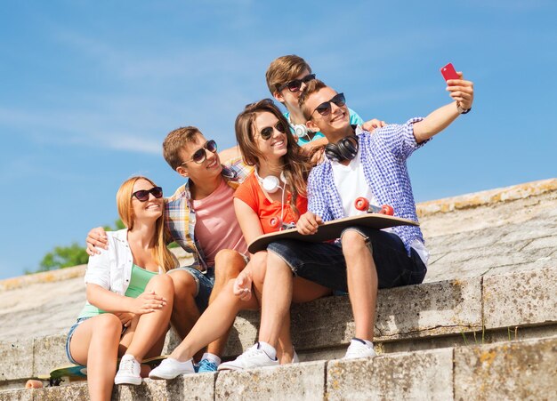 Konzept Freundschaft, Freizeit, Sommer, Technologie und Menschen - Gruppe lächelnder Freunde mit Skateboard und Smartphone, die sich im Freien selbst machen