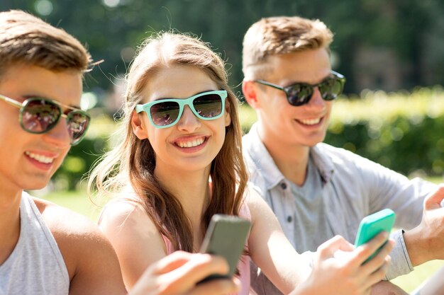 Konzept Freundschaft, Freizeit, Sommer, Technologie und Menschen - Gruppe lächelnder Freunde mit Smartphones, die auf Gras im Park sitzen