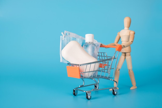 Konzept für den Kauf von Hygieneprodukten. Foto einer männlichen Holzfigur, die einen Spielzeug-Miniatur-Einkaufswagen voller Seifenmaske und Desinfektionsflüssigkeit trägt, isoliert auf blauem Hintergrund