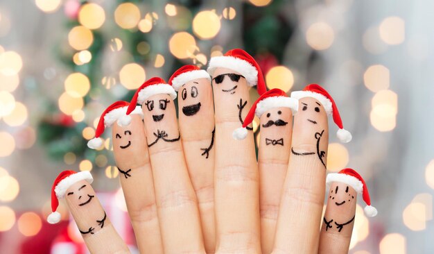 Konzept für Familie, Feiertage, Weihnachten und Körperteile - Nahaufnahme von Fingern mit Smiley-Gesichtern und Weihnachtsmützen vor hellem Hintergrund