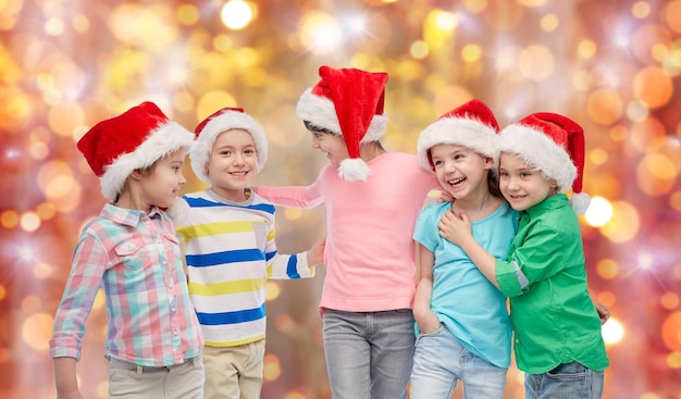Konzept für Kindheit, Weihnachten, Feiertage, Freundschaft und Menschen – eine Gruppe glücklich lächelnder kleiner Kinder in Weihnachtsmützen, die sich über Feiertagen umarmen, beleuchtet den Hintergrund