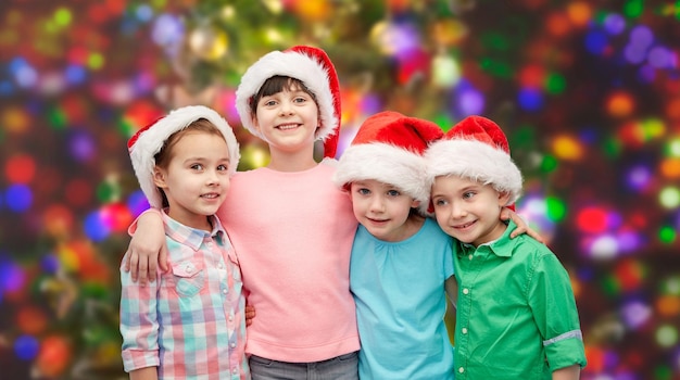 Konzept für Kindheit, Weihnachten, Feiertage, Freundschaft und Menschen – eine Gruppe glücklich lächelnder kleiner Kinder in Weihnachtsmützen, die sich über Feiertagen umarmen, beleuchtet den Hintergrund