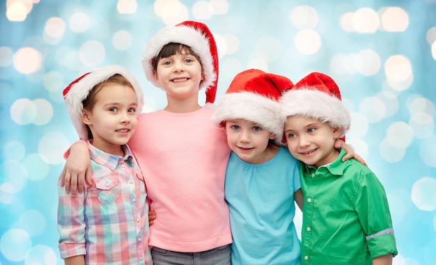 Konzept für Kindheit, Weihnachten, Feiertage, Freundschaft und Menschen - eine Gruppe glücklich lächelnder kleiner Kinder in Weihnachtsmützen, die sich vor blauem Hintergrund mit Feiertagslichtern umarmen