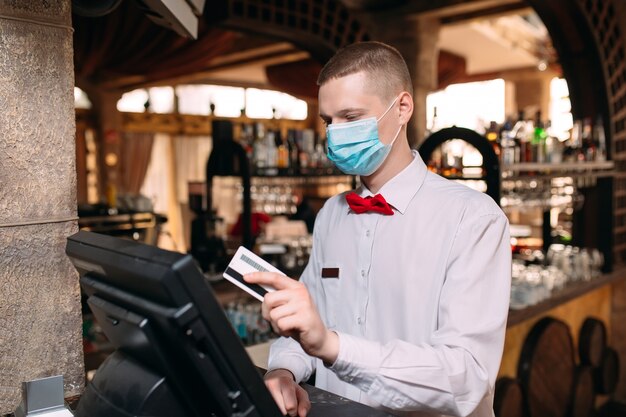 Konzept für Kleinunternehmen, Menschen und Dienstleistungen. Mann oder Kellner in der medizinischen Maske am Schalter mit Kasse, die an der Bar oder im Café arbeitet.