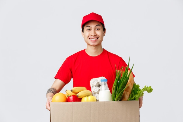 Konzept für online-shopping, lebensmittellieferung und internet-shops. fröhlich lächelnder asiatischer kerl in roter mütze und t-shirt, der eine kiste mit frischen lebensmitteln hält, stehende kundentür, mitarbeiter liefert produkt