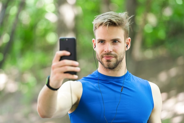 Konzept für Sportgeräte. Sportler-Handy erstellt Playlist vor dem Laufen. Mann-Athlet beschäftigtes Gesicht, das Smartphone-App, Naturhintergrund einrichtet. Sportlertraining mit Schrittzähler und Kopfhörern.