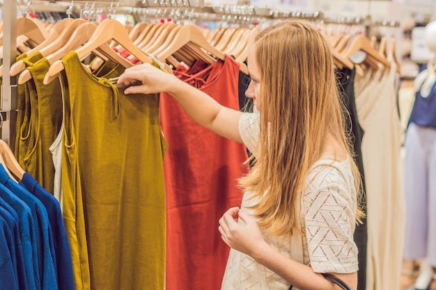 Konzept für Verkauf, Mode, Konsum und Menschen - glückliche junge Frau mit Einkaufstaschen, die Kleidung im Einkaufszentrum oder Bekleidungsgeschäft auswählen
