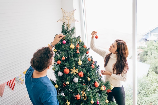 Konzept für Weihnachtspaare Glückliches junges Paar, das Pullover trägt und Spaß daran hat, verschiedene Kugeln am Weihnachtsbaum im Wohnzimmer zu Hause zu schmücken und die Weihnachtsfeiertage zu genießen und zu feiern
