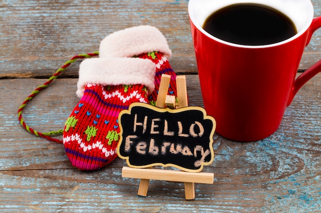 Konzept HALLO FEBRUAR Nachricht an der Tafel mit einer Tasse Kaffee und Fäustlinge