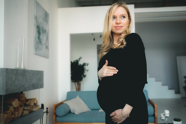 Konzept - junges schönes glückliches lächelndes schwangeres kaukasisches blondes Mädchen, das im weißen hellen Wohnzimmer steht