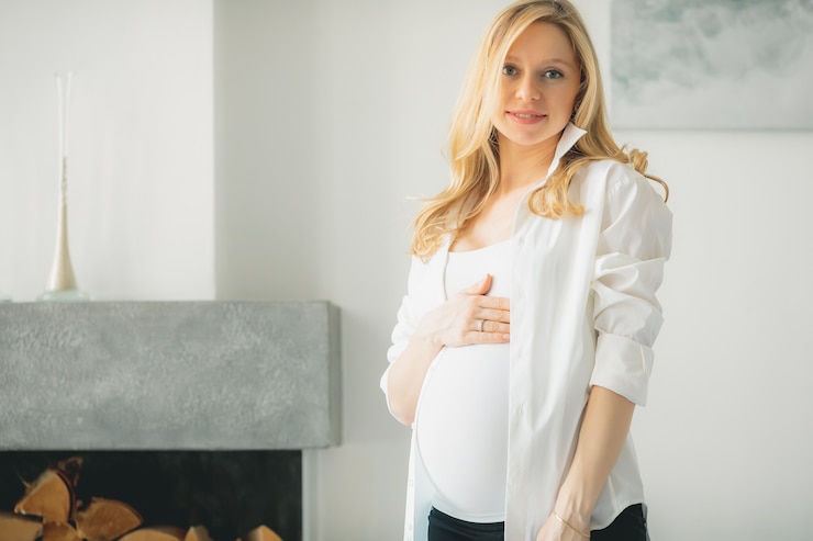 Foto konzept - junges schönes glückliches lächelndes schwangeres kaukasisches blondes mädchen, das im weißen hellen wohnzimmer steht