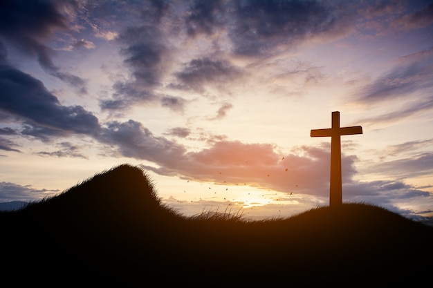 Konzept konzeptionelle schwarze Kreuzreligionssymbolschattenbild im Gras über Sonnenuntergang oder Sonnenaufganghimmel