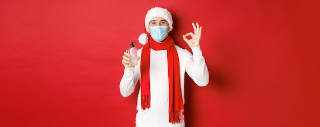 Konzept von Covid-19, Weihnachten und Feiertagen während der Pandemie. Attraktiver Mann mit Weihnachtsmütze und medizinischer Maske, der ein Okay-Zeichen zeigt, während er Händedesinfektionsmittel empfiehlt und auf rotem Hintergrund steht.