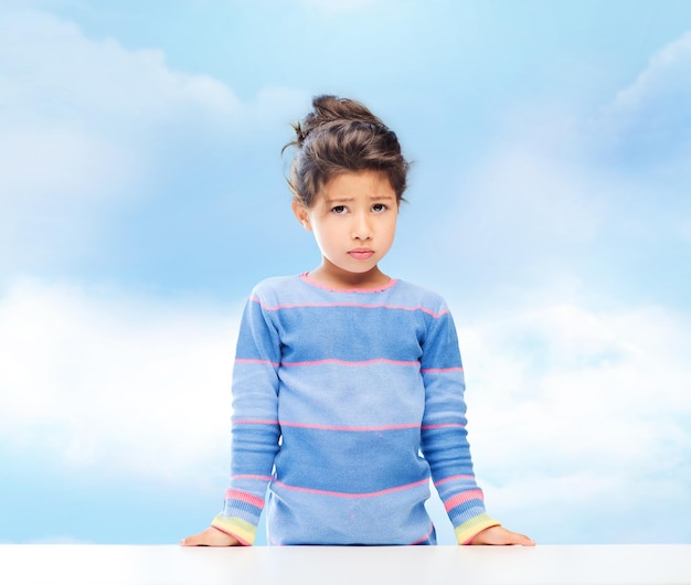 Konzept von Menschen, Kindheit und Emotionen - trauriges kleines Mädchen über blauem Himmelshintergrund