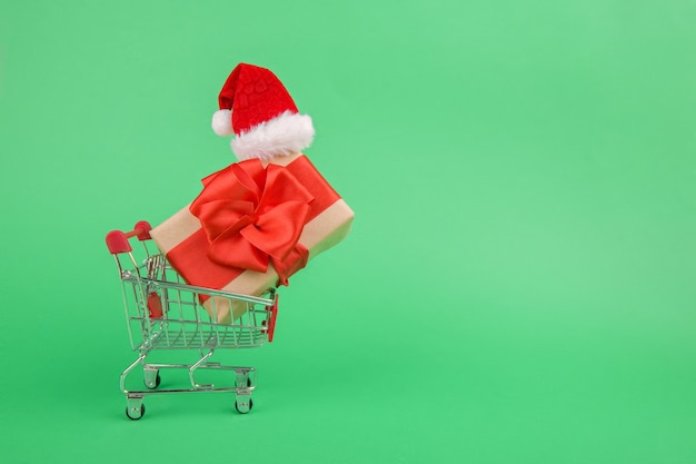 Konzeptionelle Online-Weihnachtseinkaufsbox mit einem Geschenk und einem grünen Band im Mini-Einkaufswagen oder im Wagen neben einem