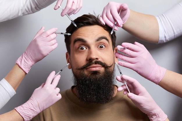 Konzeptionelles Schönheits- und Kosmetologiebild der Hände mehrerer Ärzte, die Spritzen über das männliche Gesicht halten